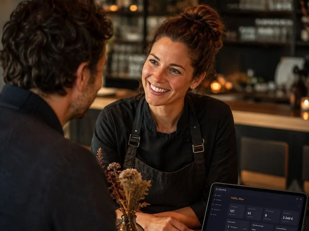 Gastronomin im persönlichen Gespräch mit einem Gast am Tresen, im Hintergrund ein Tablet mit dem EATRIQ-Dashboard
